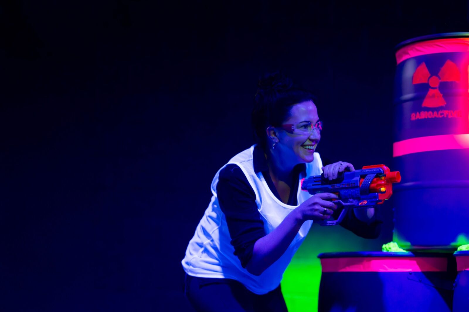 Amanda, co-owner of Blast'D! Arena, smiling mid-game while aiming a foam blaster beside a glowing barrel