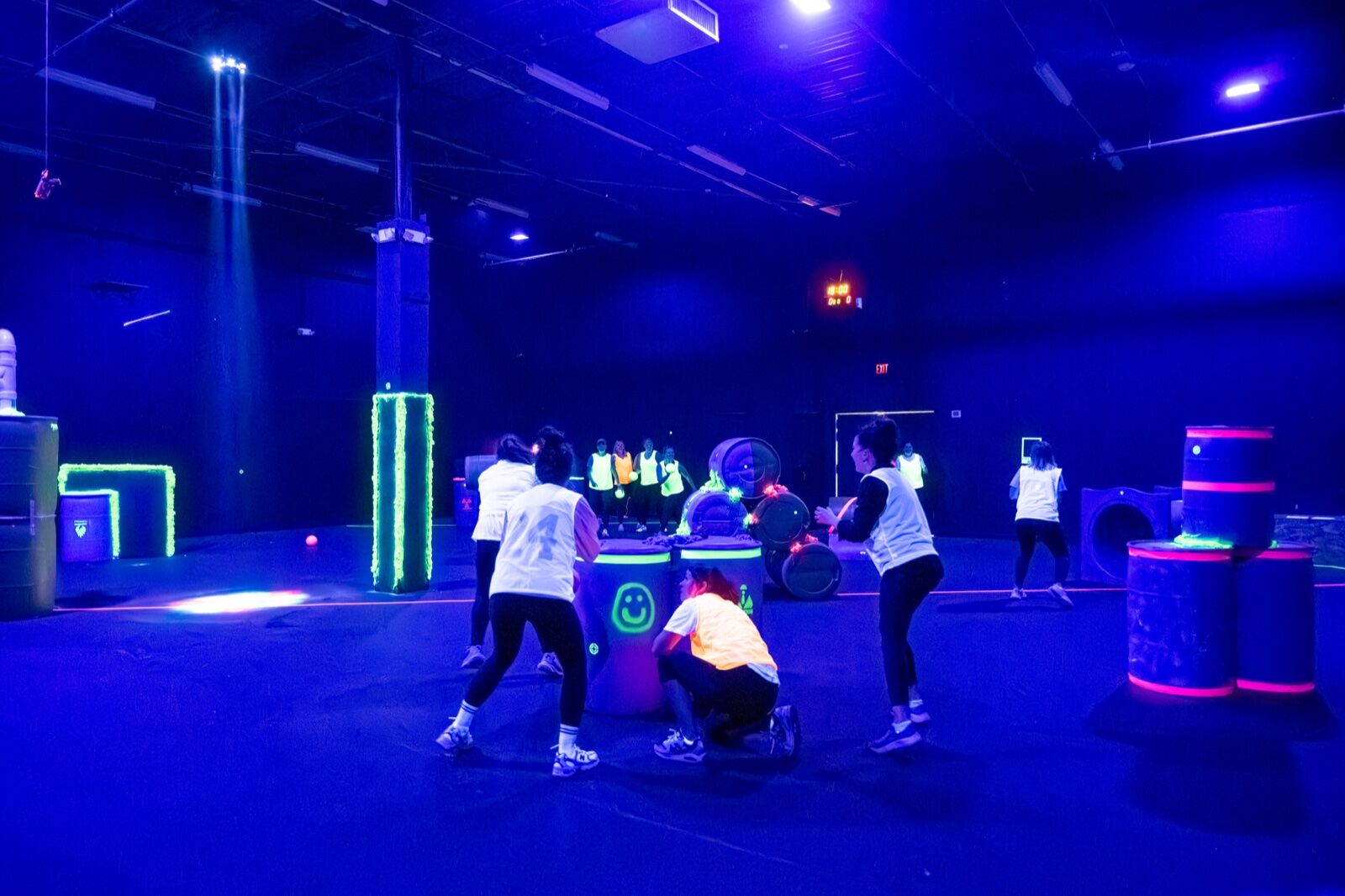 Players spread across the blacklit Blast'D! arena floor mid-game, with glowing barrel bunkers and a spotlight beam