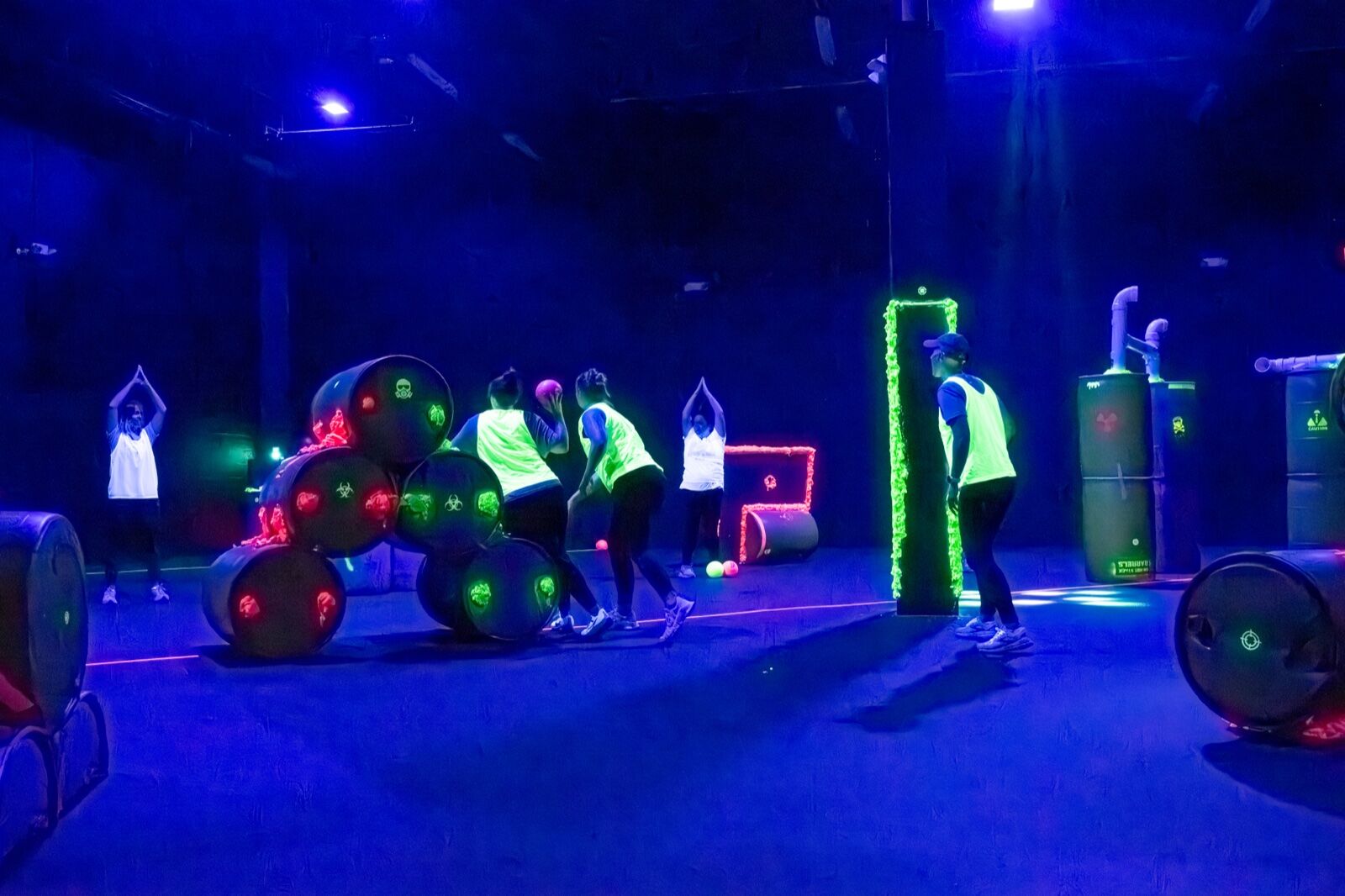 A wide view of the blacklit arena mid-game with barrel cover and players in motion
