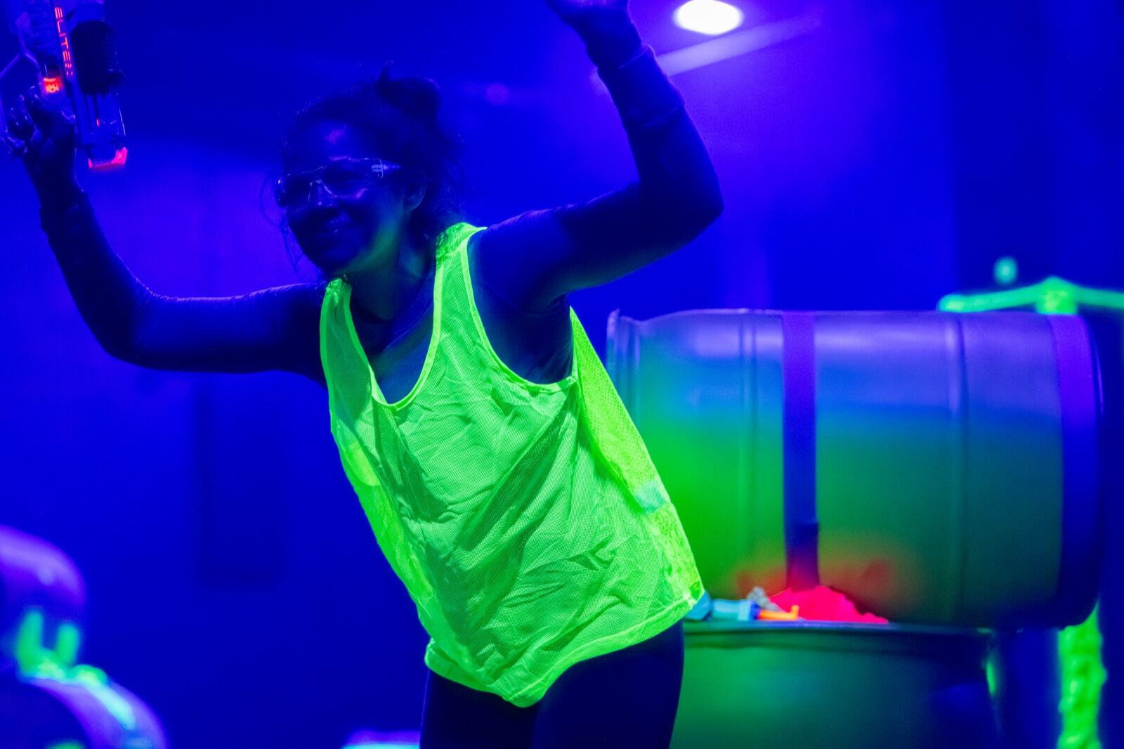 A smiling player in a glowing neon vest and safety glasses raising a foam blaster under the blacklights