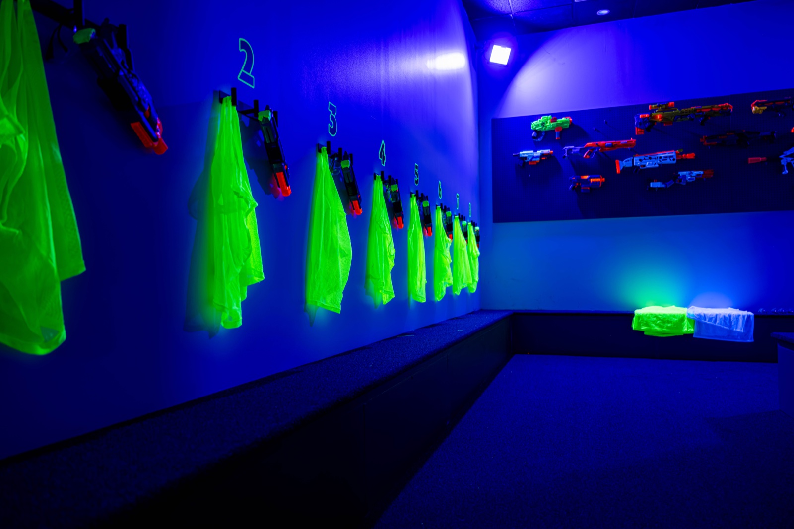 Numbered hooks of glowing neon jerseys and foam blasters along the armory wall under blacklight