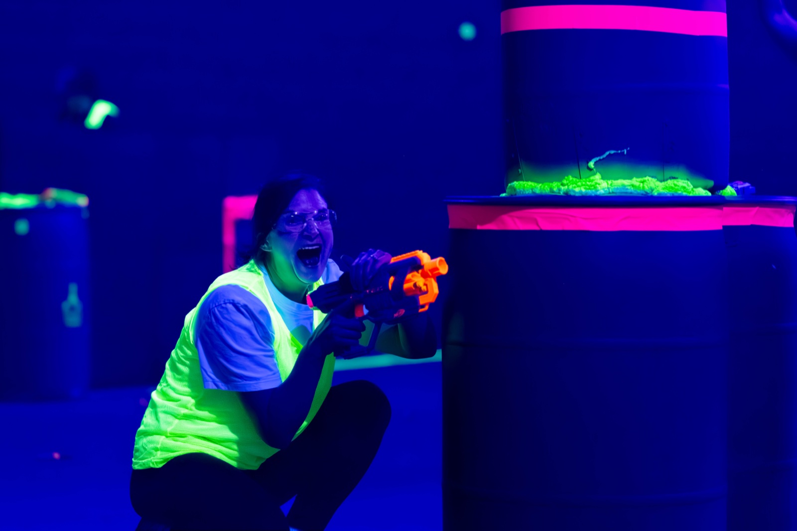A player in a neon vest and safety glasses laughing while aiming a foam blaster in the blacklit arena